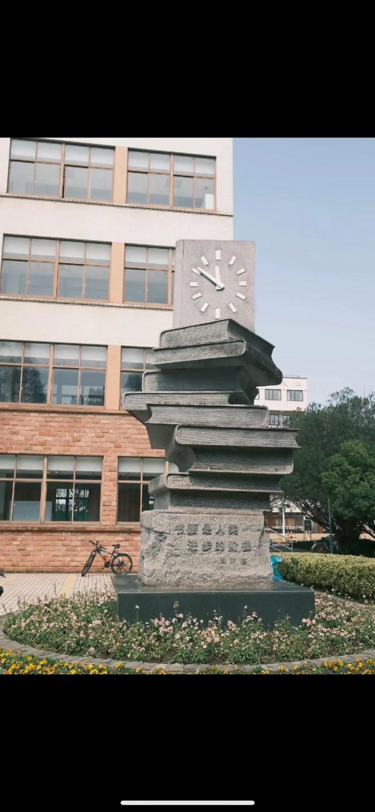 A stone monument or clock sculpture stands in a landscaped plaza in front of a modern building with large windows and trees under a blue sky.