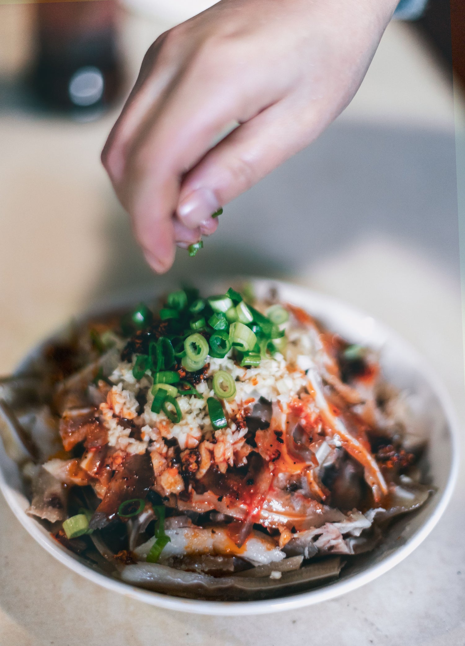 A hand scatters chopped scallions over a spicy Sichuan dish.