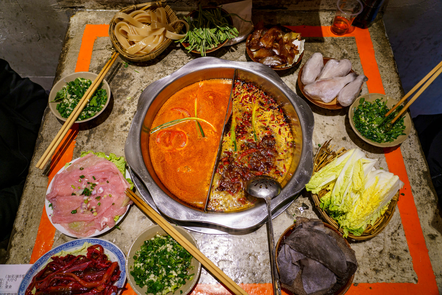 Divided spicy Chongqing hotpot surrounded by sliced meat, vegetables, herbs and sauces.
