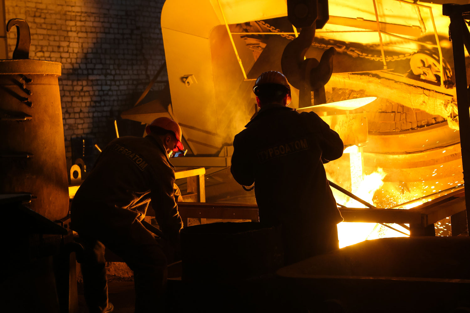 Two helmeted steelworkers seen from behind beside a glowing molten-metal furnace.