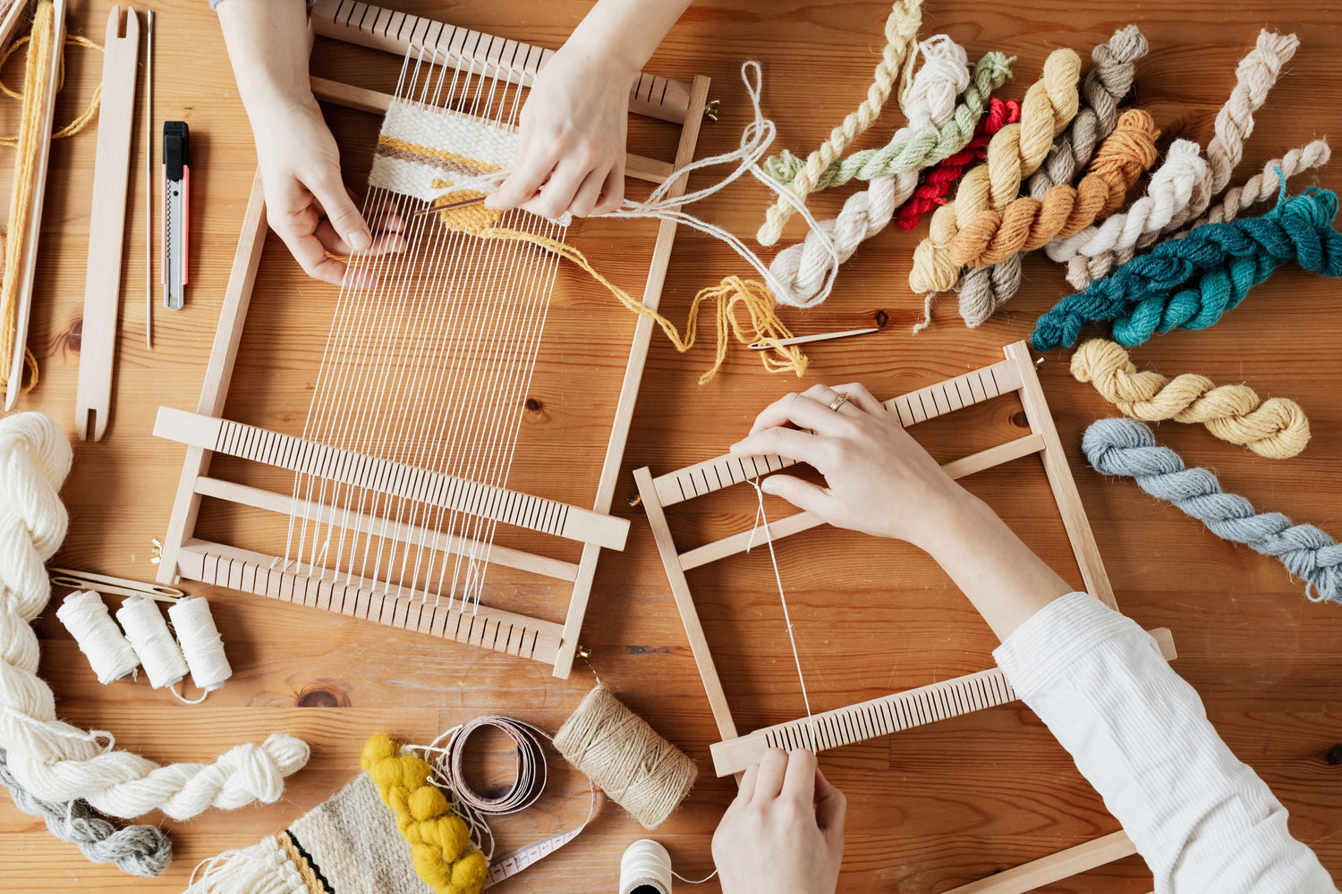 Two pairs of hands weaving natural-coloured yarn on small wooden looms at a workshop table.