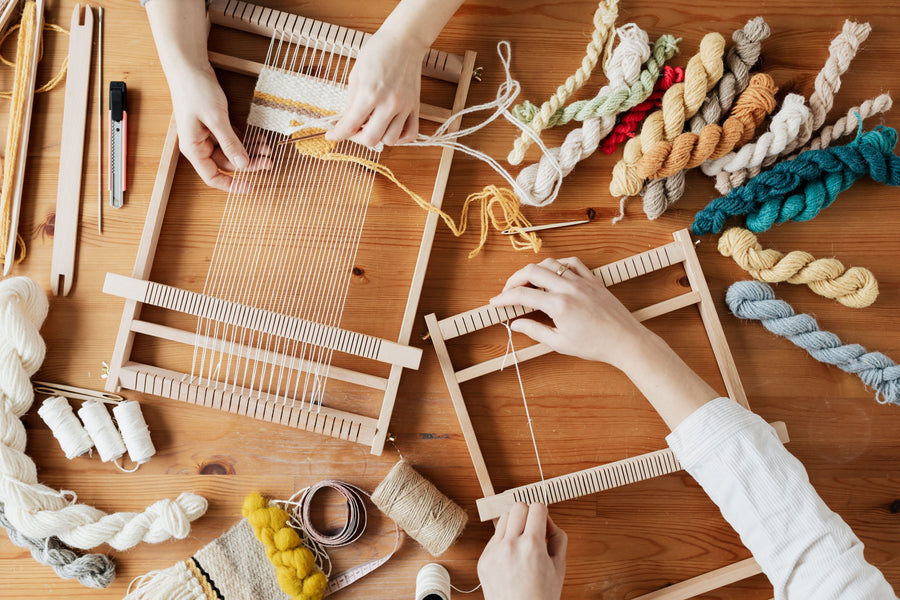 Two pairs of hands weaving natural-coloured yarn on small wooden looms at a workshop table.
