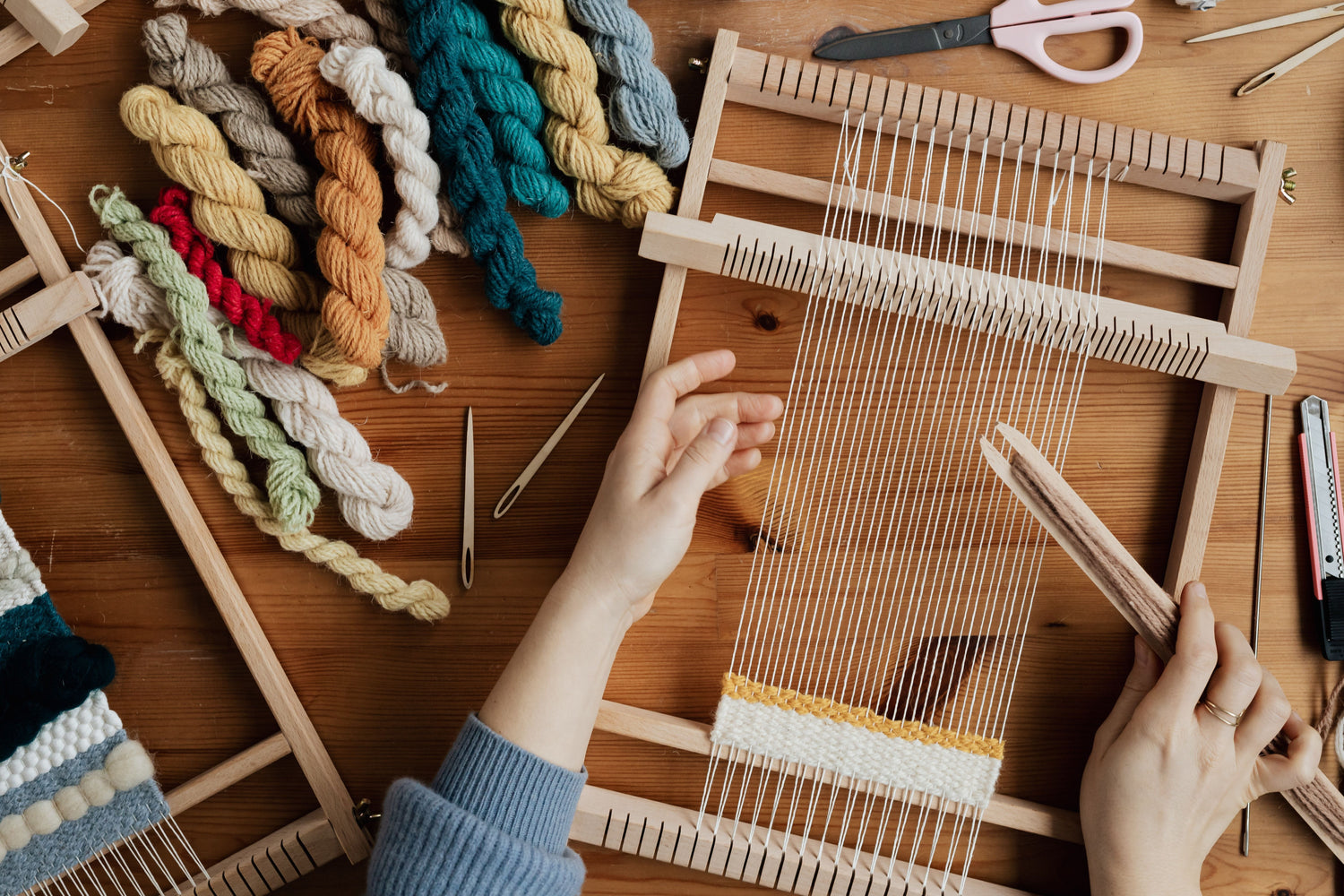 Hands weaving cream and mustard yarn on a small wooden hand loom.