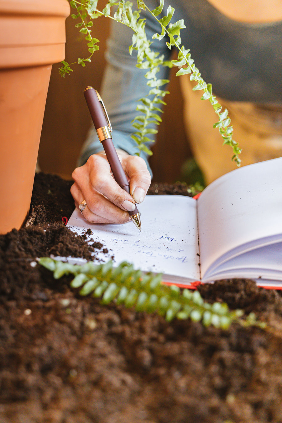 Hand writing field notes in an open notebook among soil and green plants.
