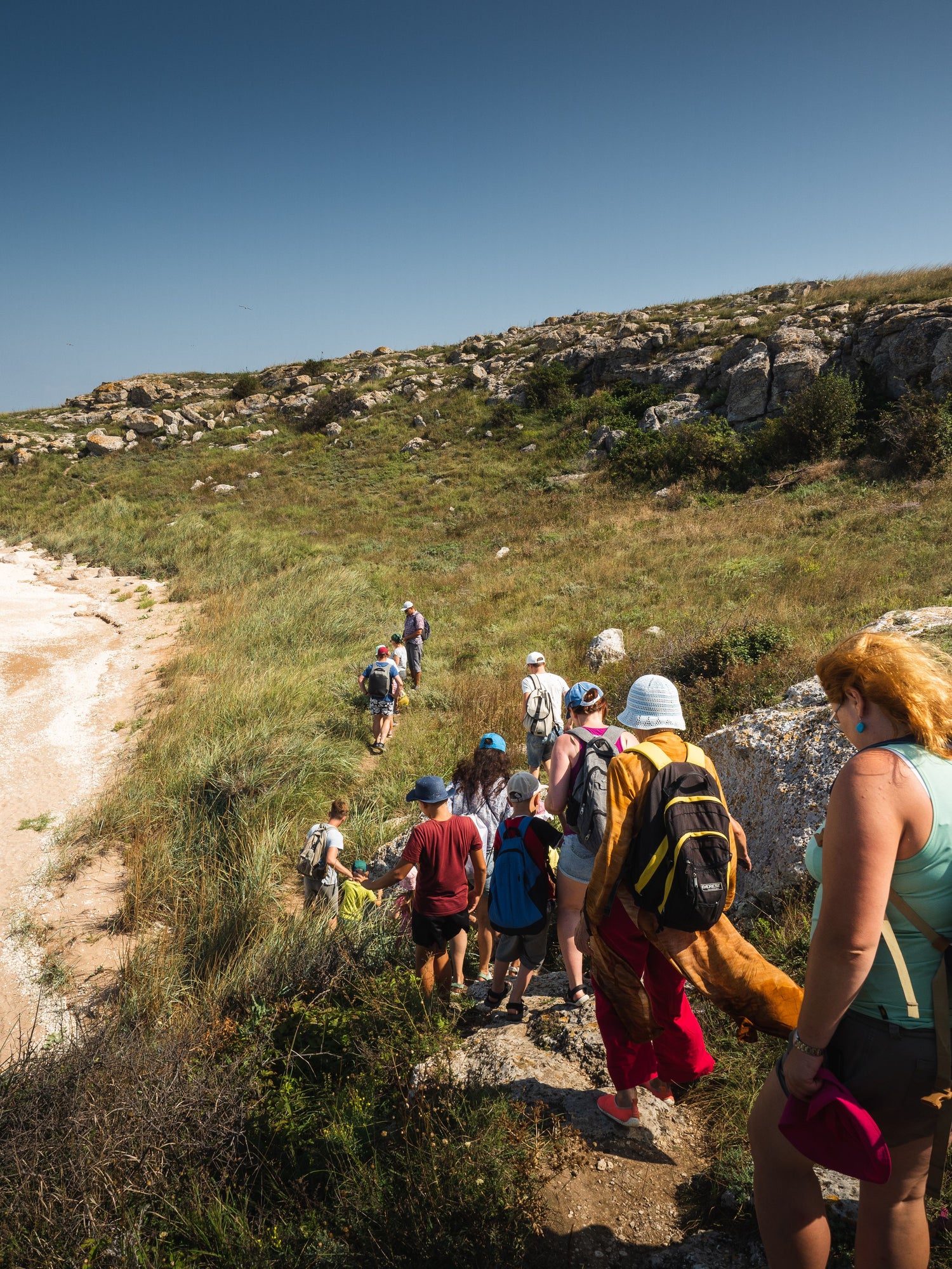 Group of hikers seen from behind following a narrow trail across a rocky hillside.