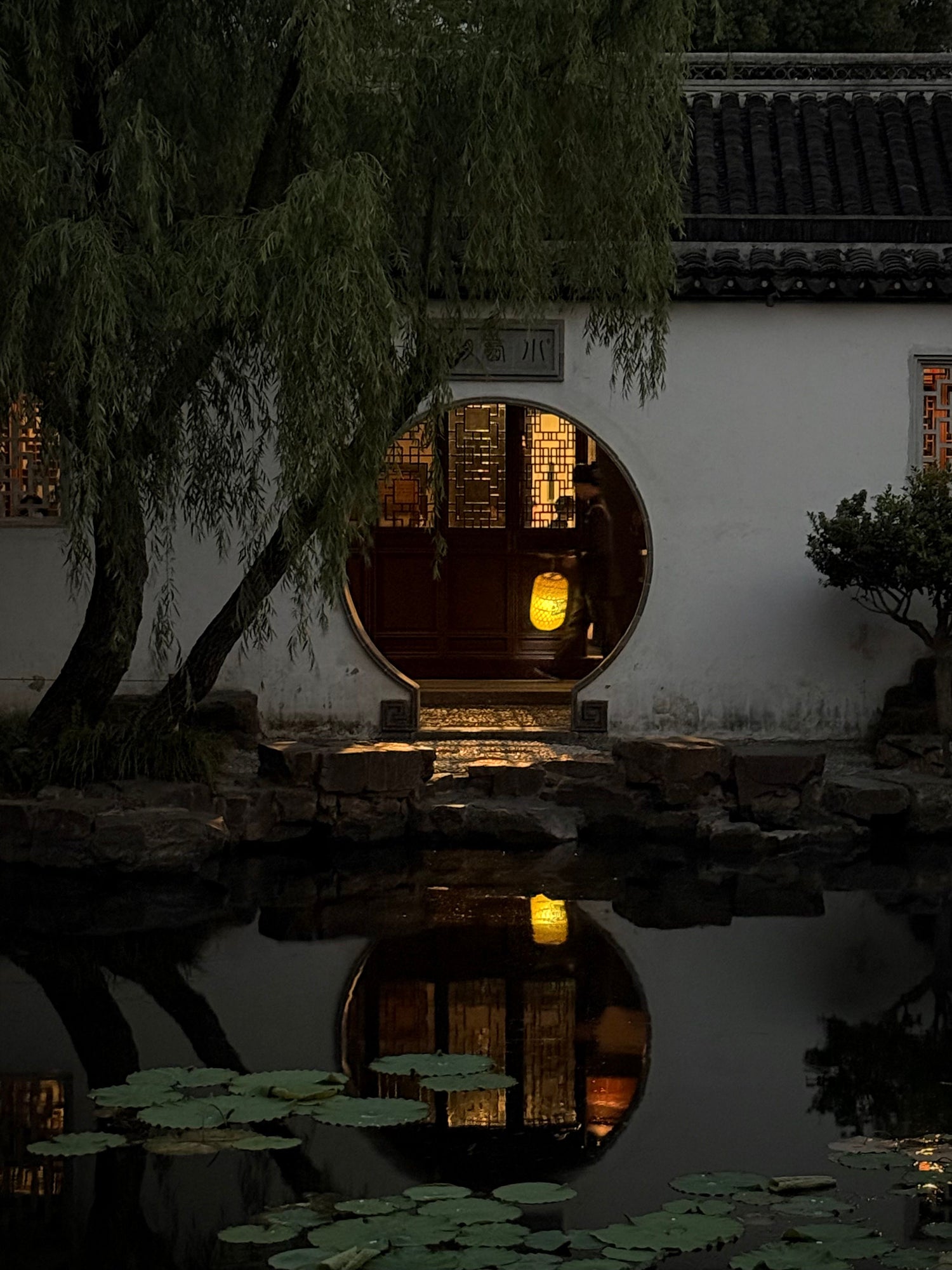 An illuminated moon gate reflected in a pond in a classical Suzhou garden at dusk.