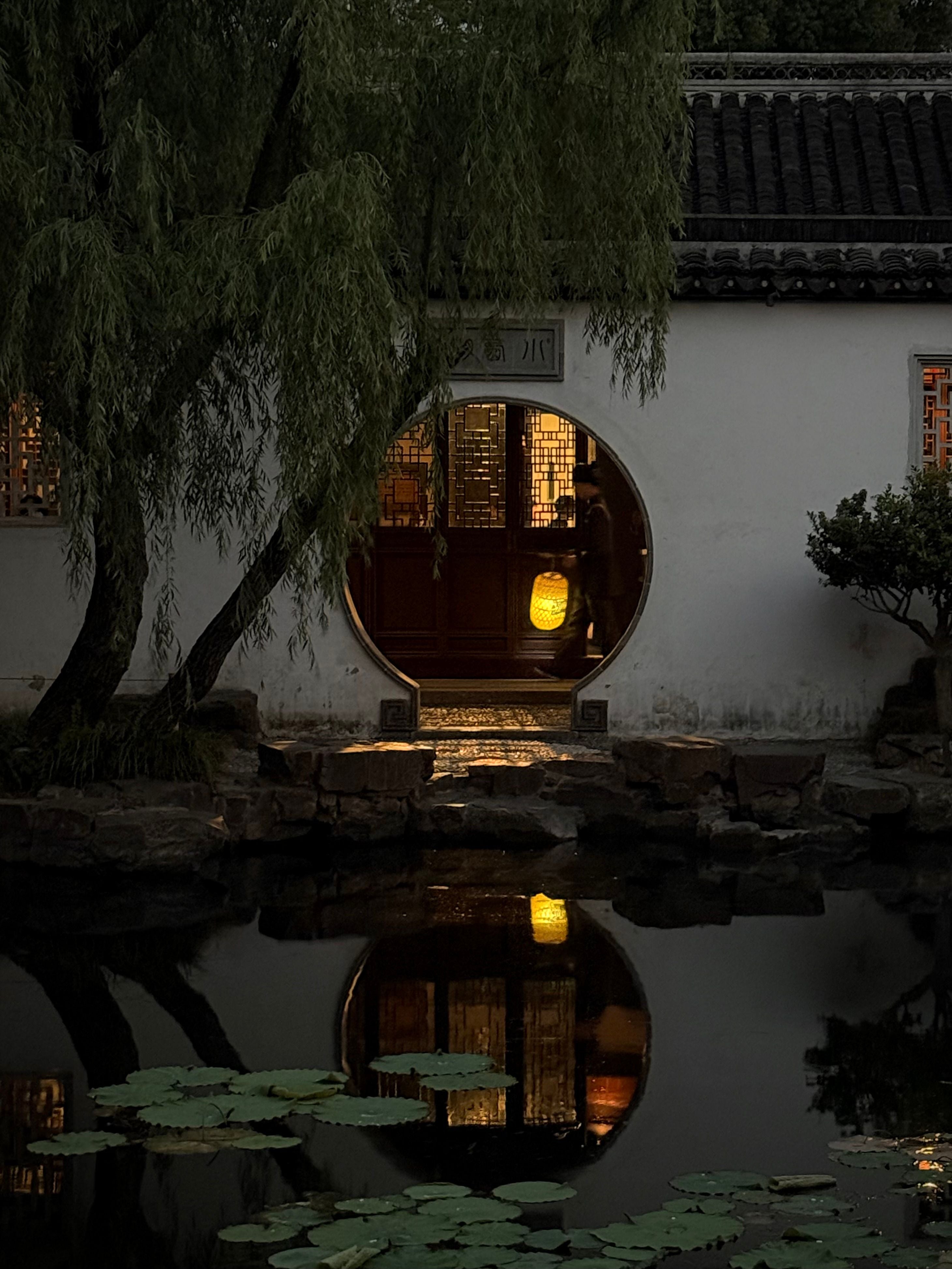 An illuminated moon gate reflected in a pond in a classical Suzhou garden at dusk.