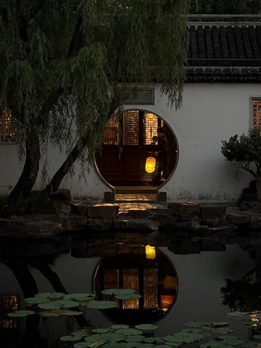 An illuminated moon gate reflected in a pond in a classical Suzhou garden at dusk.